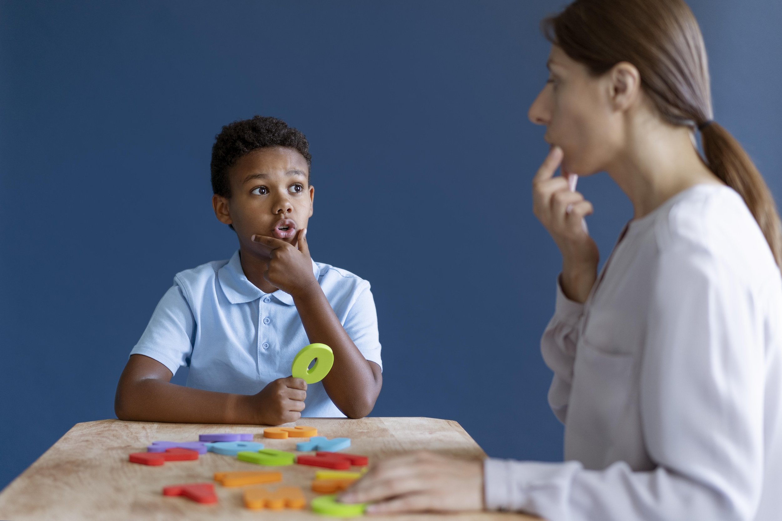 A young boy with curly hair and a blue shirt is sitting at a wooden table, holding colorful letter shapes and appears to be in a conversation with a woman. The woman has long red hair tied back and is wearing a white top. She is touching her chin and listening attentively. There are scattered colorful letter shapes on the table, and the background is solid blue.