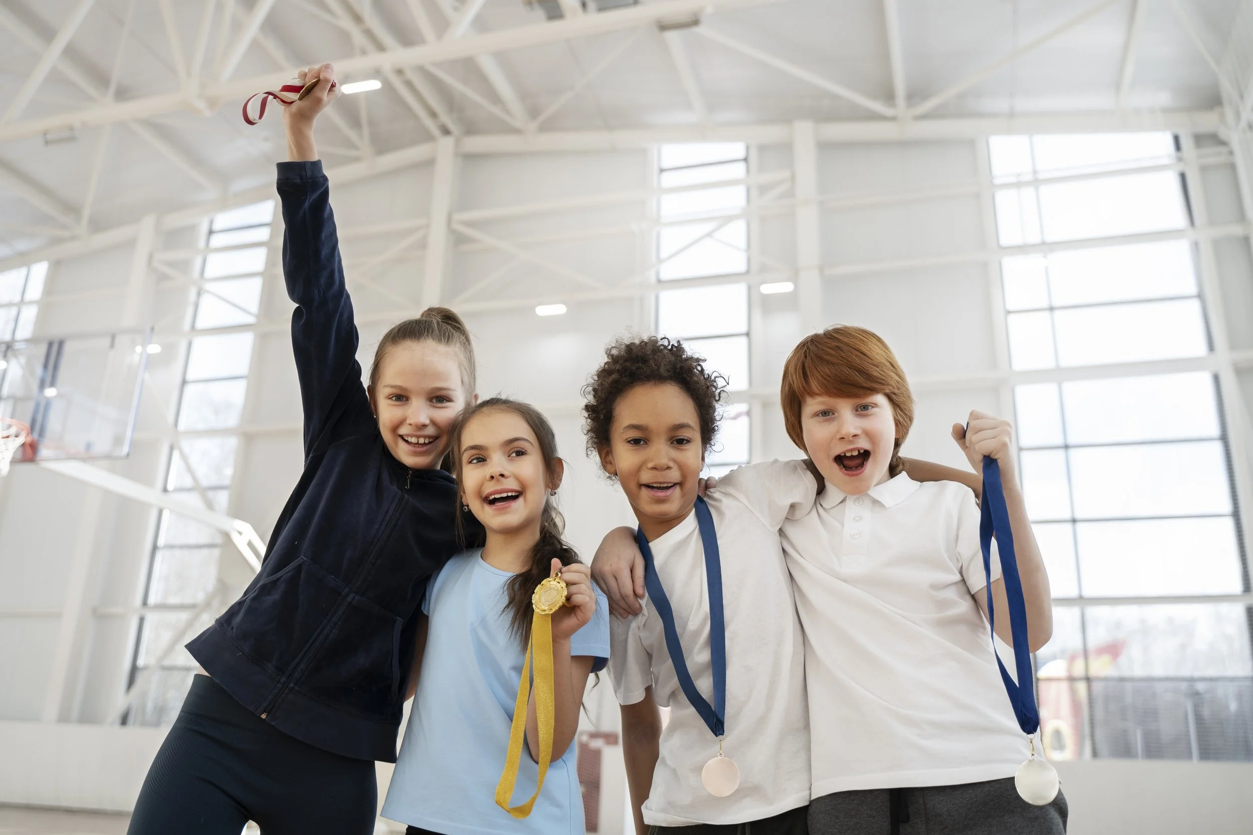 A group of five children standing together in a gymnasium, smiling and showing medals around their necks, with one child raising a trophy with a ribbon in the air.