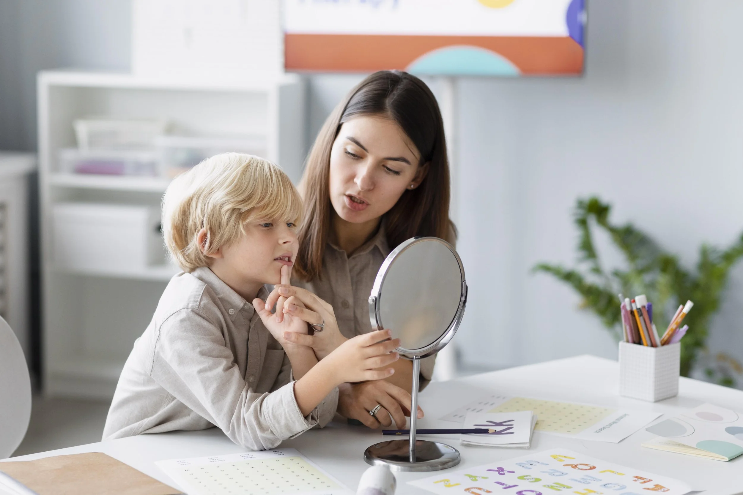 A woman and a young boy sitting at a desk, looking into a round mirror, surrounded by papers and colored pens, with a background of a modern room with artwork and plants.