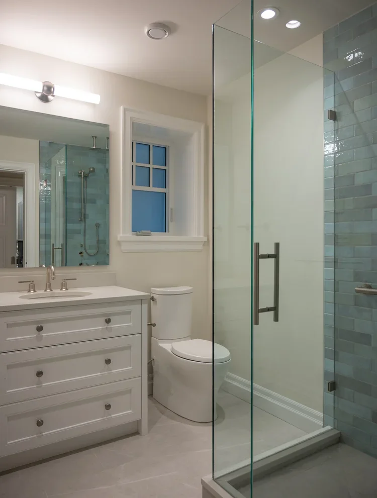 Modern bathroom with white vanity, mirror, toilet, and glass-enclosed shower with blue tiles.