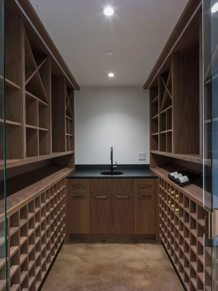A walk-in wine cellar with wooden shelves and cubbies for wine bottles, a black countertop with a sink, and a small window or glass door on the right.