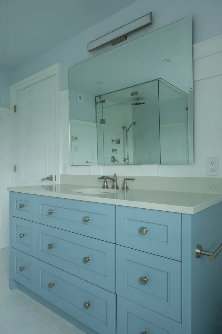 Blue bathroom vanity with multiple drawers and a white countertop, with a large mirror and a visible glass-enclosed shower in the reflection.