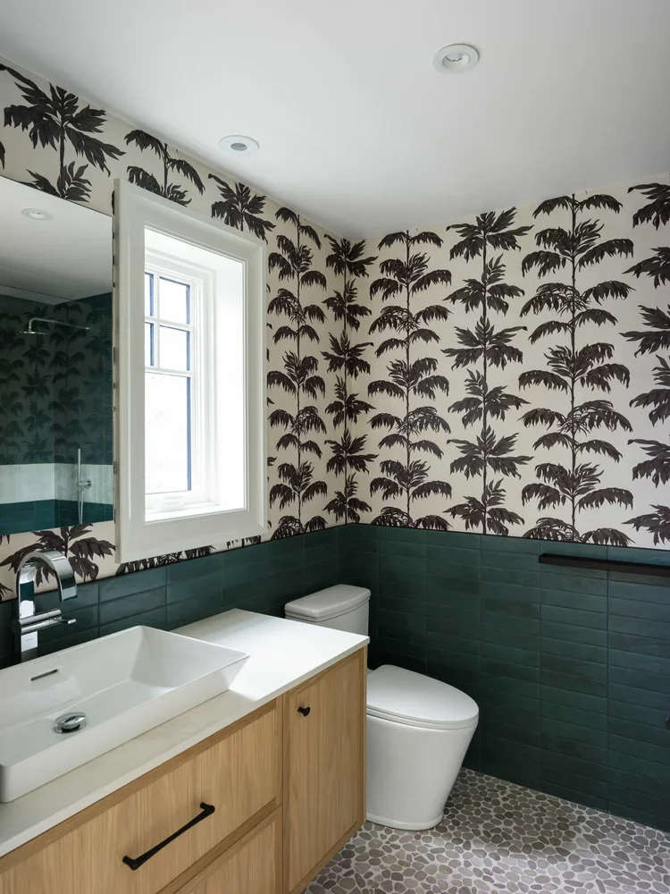 Bathroom with tropical palm tree patterned wallpaper, green tiled wainscoting, wooden vanity with a white rectangular vessel sink, and a modern white toilet. There is a window letting in natural light and a large mirror on the wall.