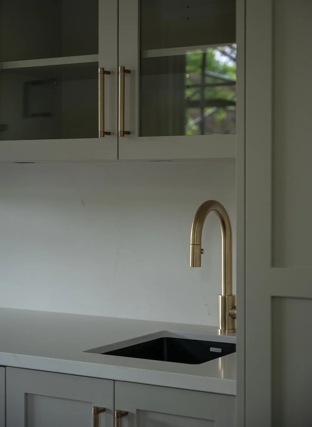 Modern kitchen with grey cabinets, gold handles, a black sink, and a gold faucet, with a window showing trees outside.