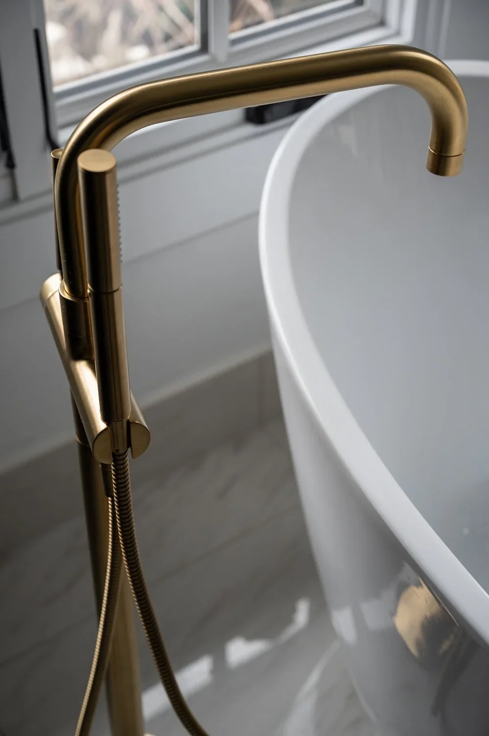 Close-up of a brass-colored freestanding bathtub faucet next to a white bathtub with a curved edge, near a window with gray blinds.