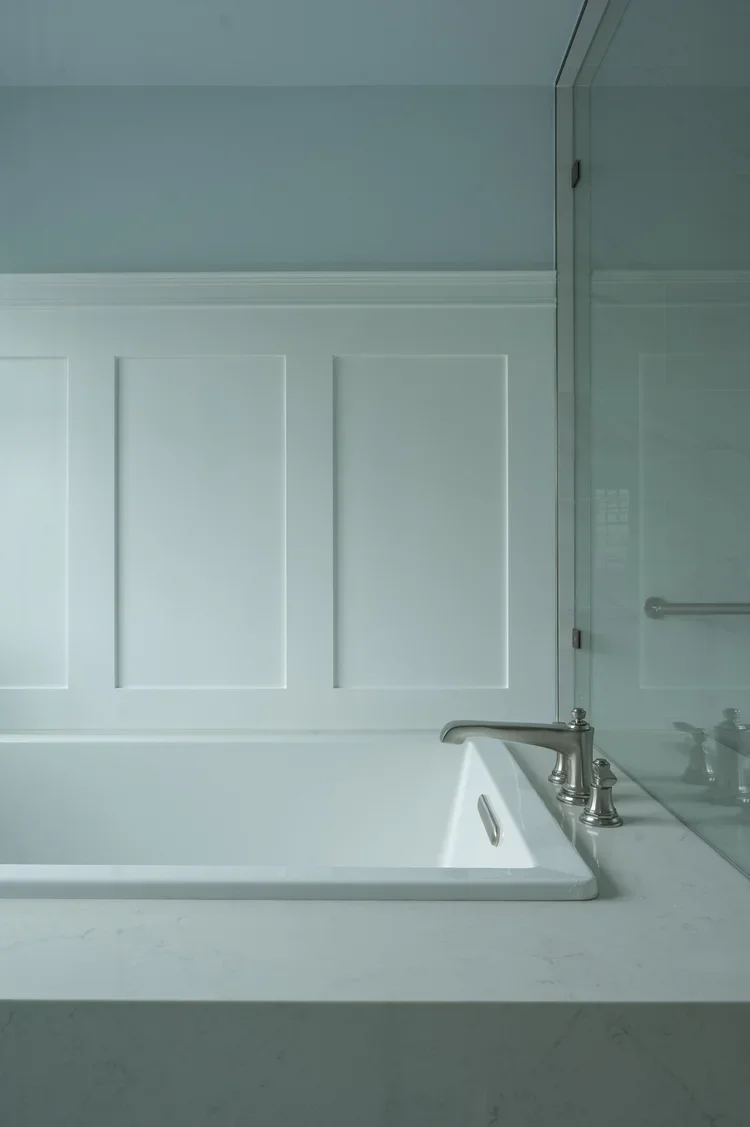 A white bathtub with silver fixtures next to a glass shower wall in a bathroom with light blue walls and white paneling.