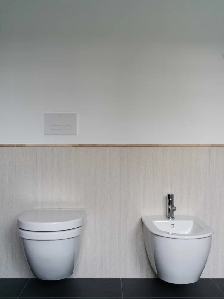 Toilet and bidet in a modern bathroom with white fixtures, dark floor tiles, and a light-colored wall.