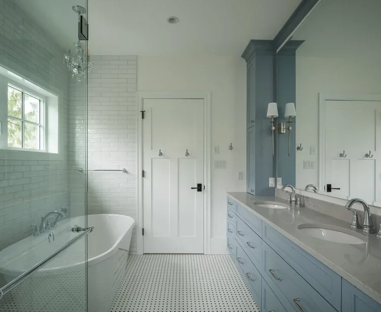 Bathroom with a bathtub, glass shower door, white tile walls, and a double sink vanity with blue cabinets, wall-mounted sconces, and a large mirror.