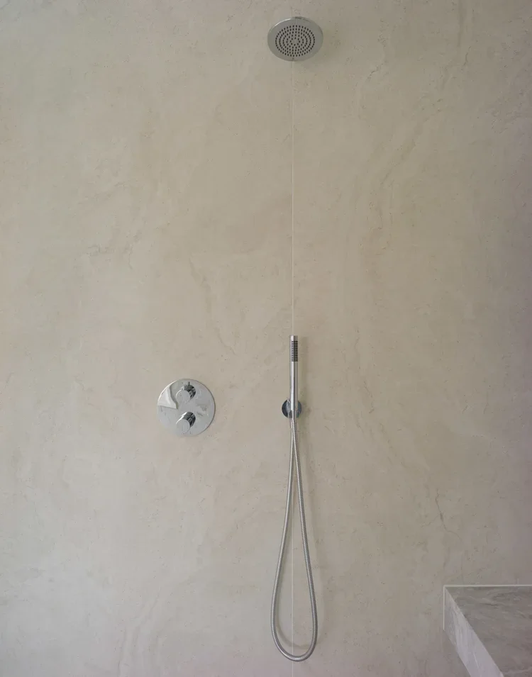 A modern shower with a ceiling-mounted showerhead, handheld shower wand, and controls on a beige tiled wall.