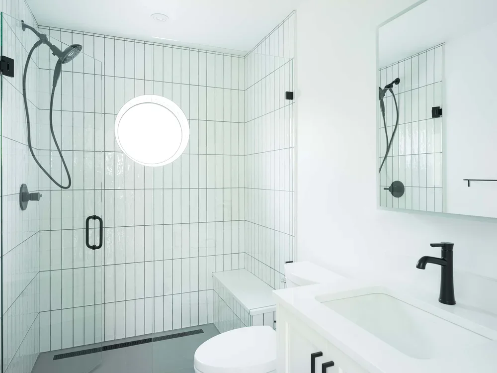 Modern minimalist bathroom with white tiled shower area, round window, white vanity with black faucet, and a large mirror.