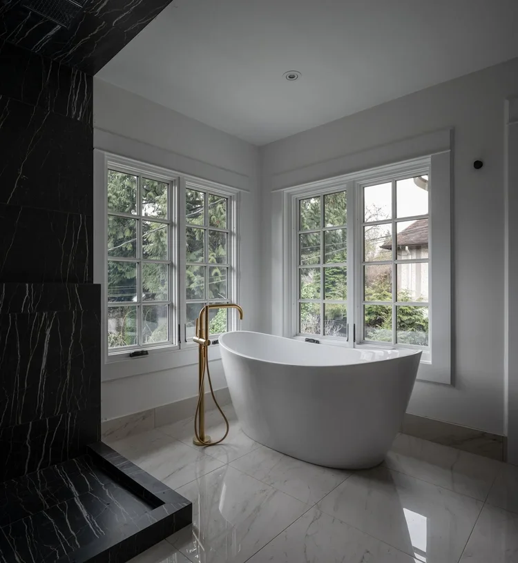 Modern bathroom featuring a standalone bathtub near three large windows with greenery outside, a gold freestanding faucet, and black marble accents on the wall.