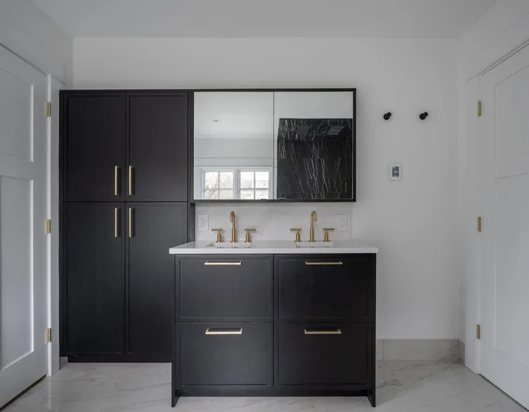 Modern bathroom vanity with black cabinetry, two gold faucets, large mirror, white countertop, and black marble accent. White walls with gold hardware on the doors.