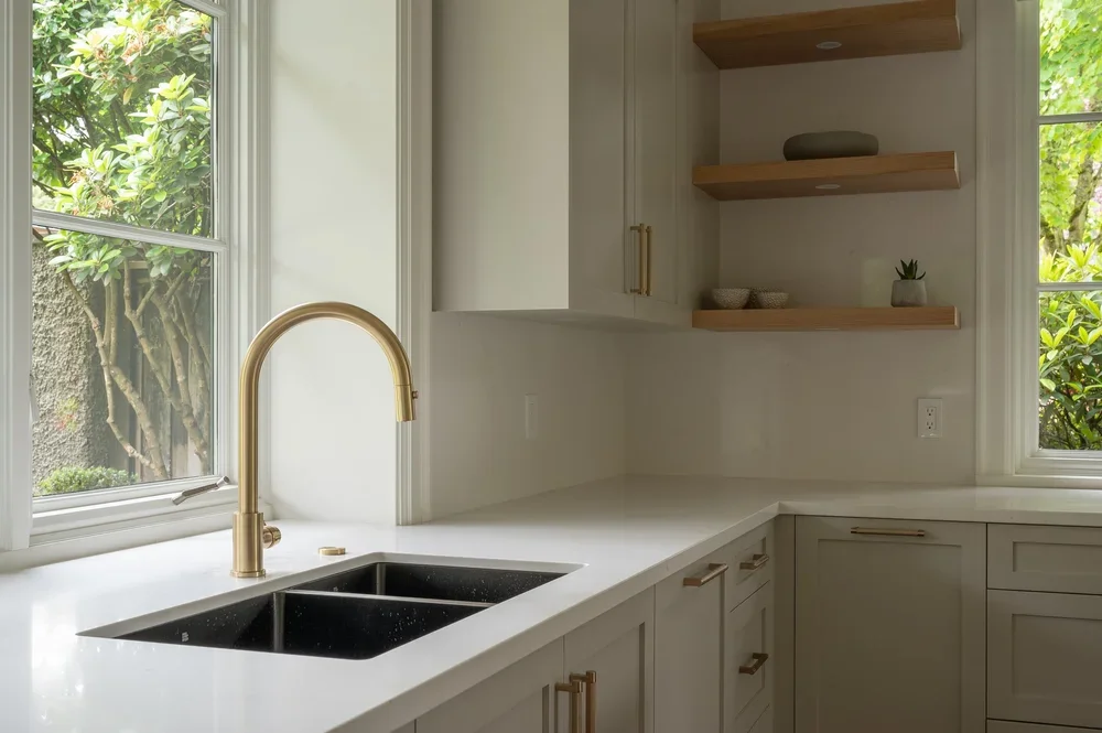 Bright kitchen with white countertops, a black double sink, and gold fixtures. Two large windows showing green trees outside, with open wooden shelves on the wall holding decorative items.