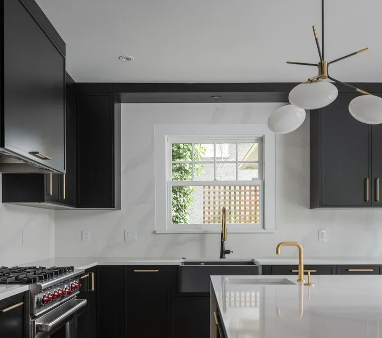 Modern kitchen with black cabinets, marble countertops, a large window, and gold accents on the faucet and cabinet handles.