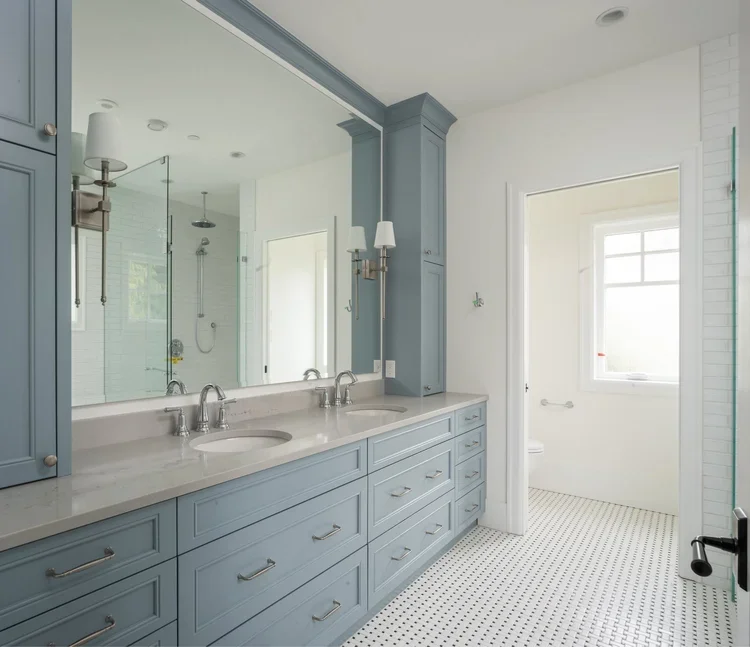 Bathroom with double vanity, blue cabinets, marble countertop, large mirror, wall sconce, glass-enclosed shower, white tile walls, window, and small black and white patterned floor.