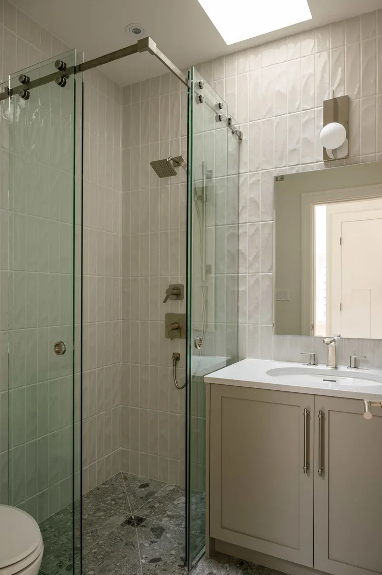A modern bathroom featuring a walk-in glass shower with a rainfall showerhead and a white vanity with a sink mirror. The ceiling has a skylight providing natural light.