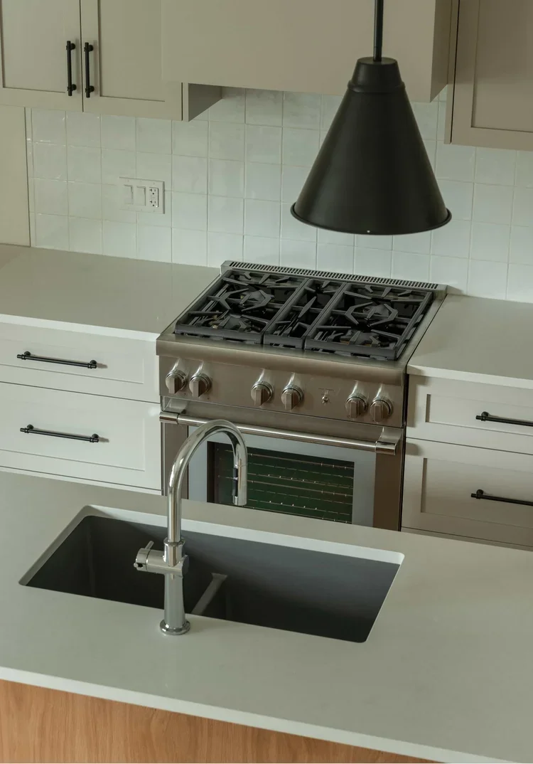 Modern kitchen with white countertops, beige cabinets, a black pendant light, a stainless steel gas stove, and a black sink with a silver faucet.