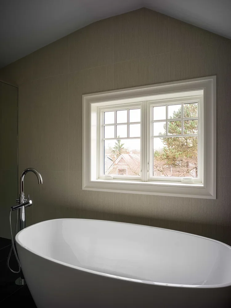 A white modern bathtub beside a window in a bathroom with beige walls and a silver floor-mounted faucet.