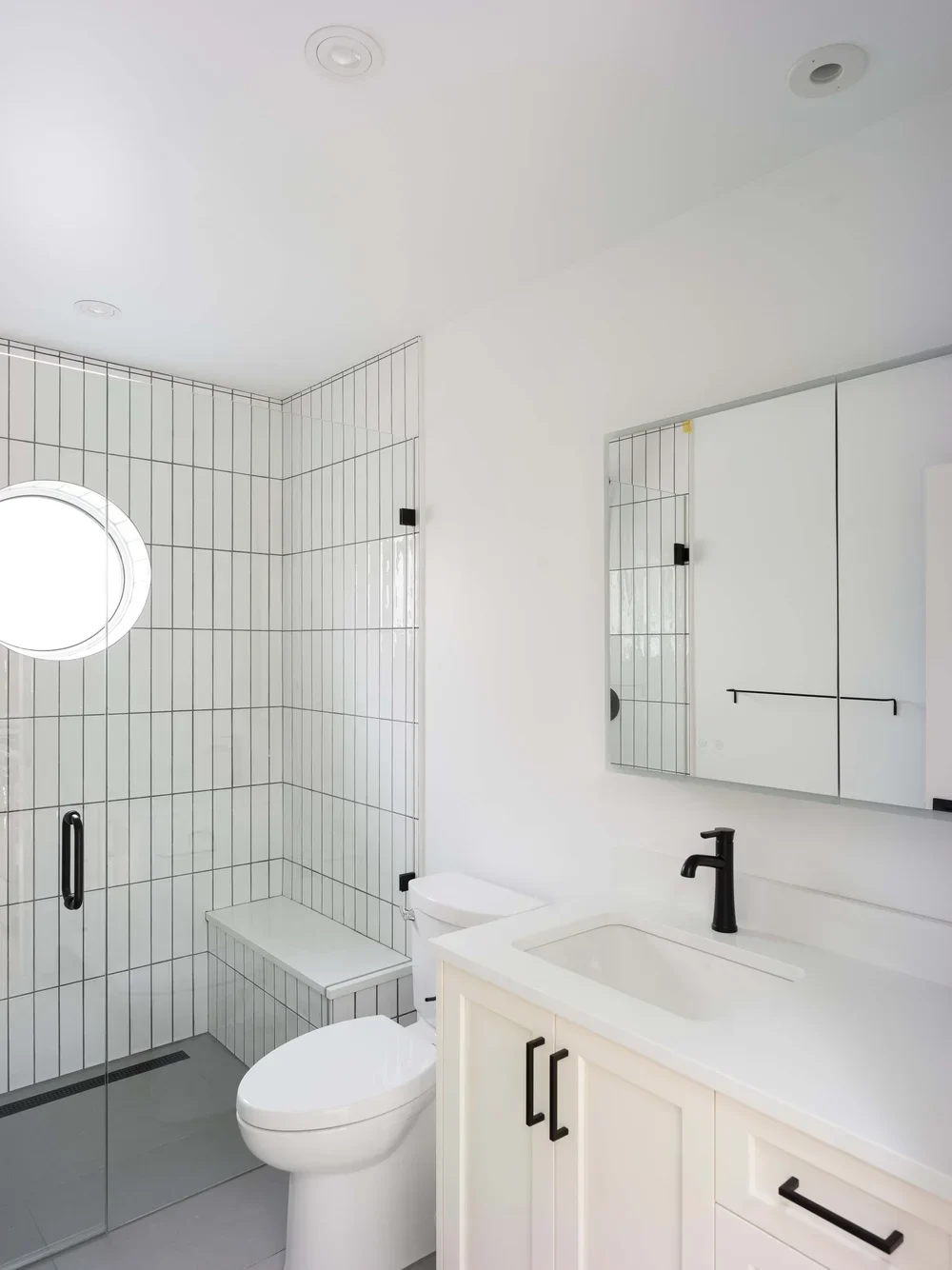 Modern white bathroom with black fixtures, including a black faucet and cabinet handles, a glass shower with white tile and black trim, a round window, and a large mirror above a white vanity.