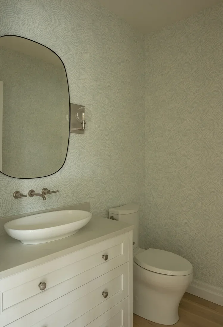 A modern bathroom with a white vanity, oval vessel sink, wall-mounted faucet, large oval mirror, wall sconce light, and a toilet, all against light-colored wallpapered walls.