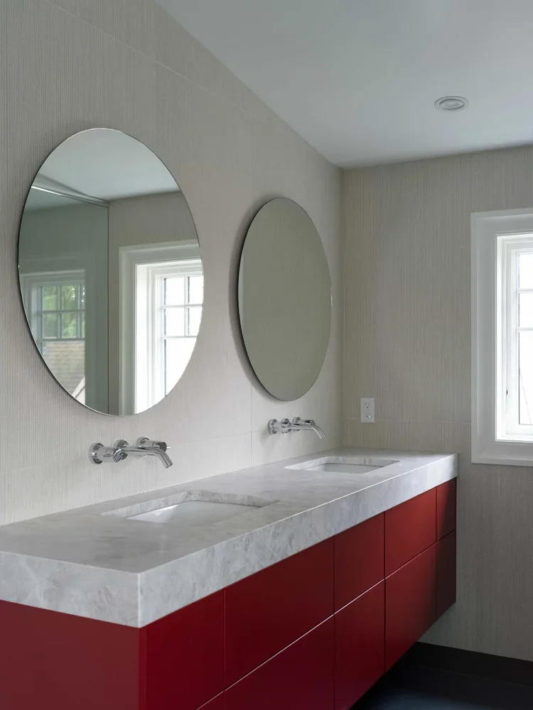 Modern bathroom with a double marble countertop sink, two round mirrors, off-white walls, a window with white trim, and a red cabinet below the sinks.
