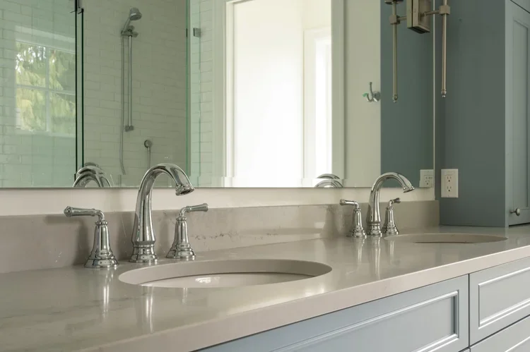 Bathroom sink with two faucets, large mirror, and a shower visible in the reflection.