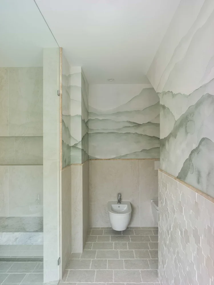 Interior of a modern bathroom with wall-mounted bidet, stone tile flooring, and a watercolor mountain mural on the walls.