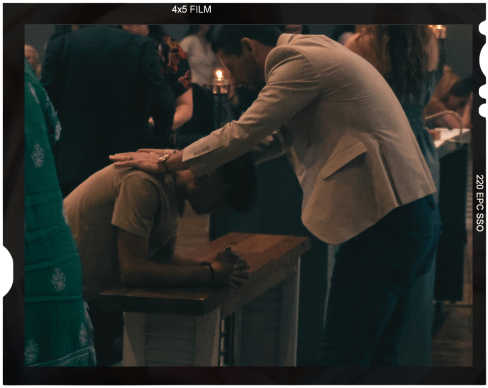 Young man in a jacket is praying over a youth male in a worship service. Both men have their heads bowed