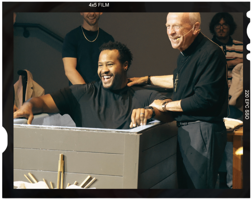 An adult male is sitting in a baptism tub smiling very big with the pastor standing next to him also smiling