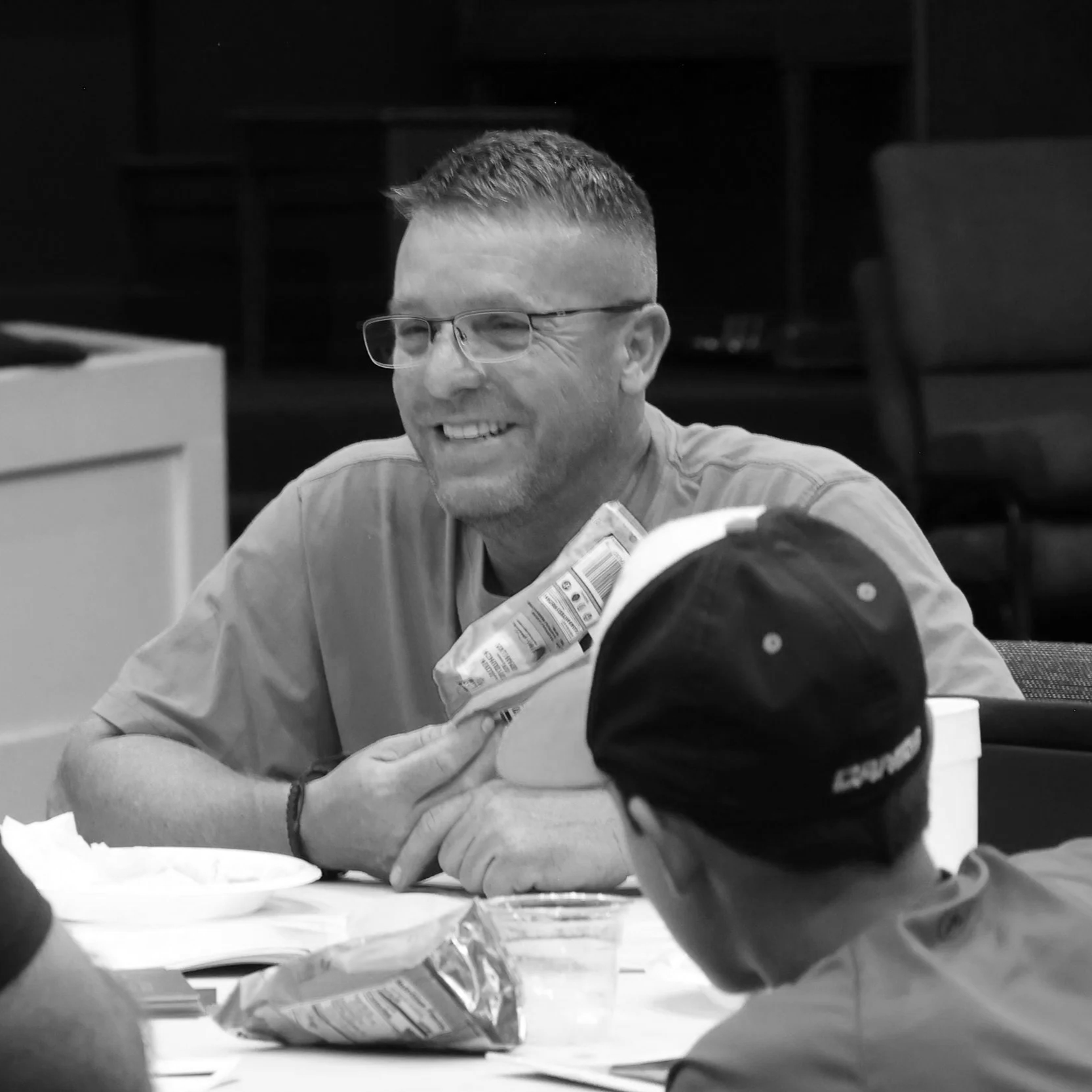 Man with glasses sitting at a table with other men. Man is smiling holding food
