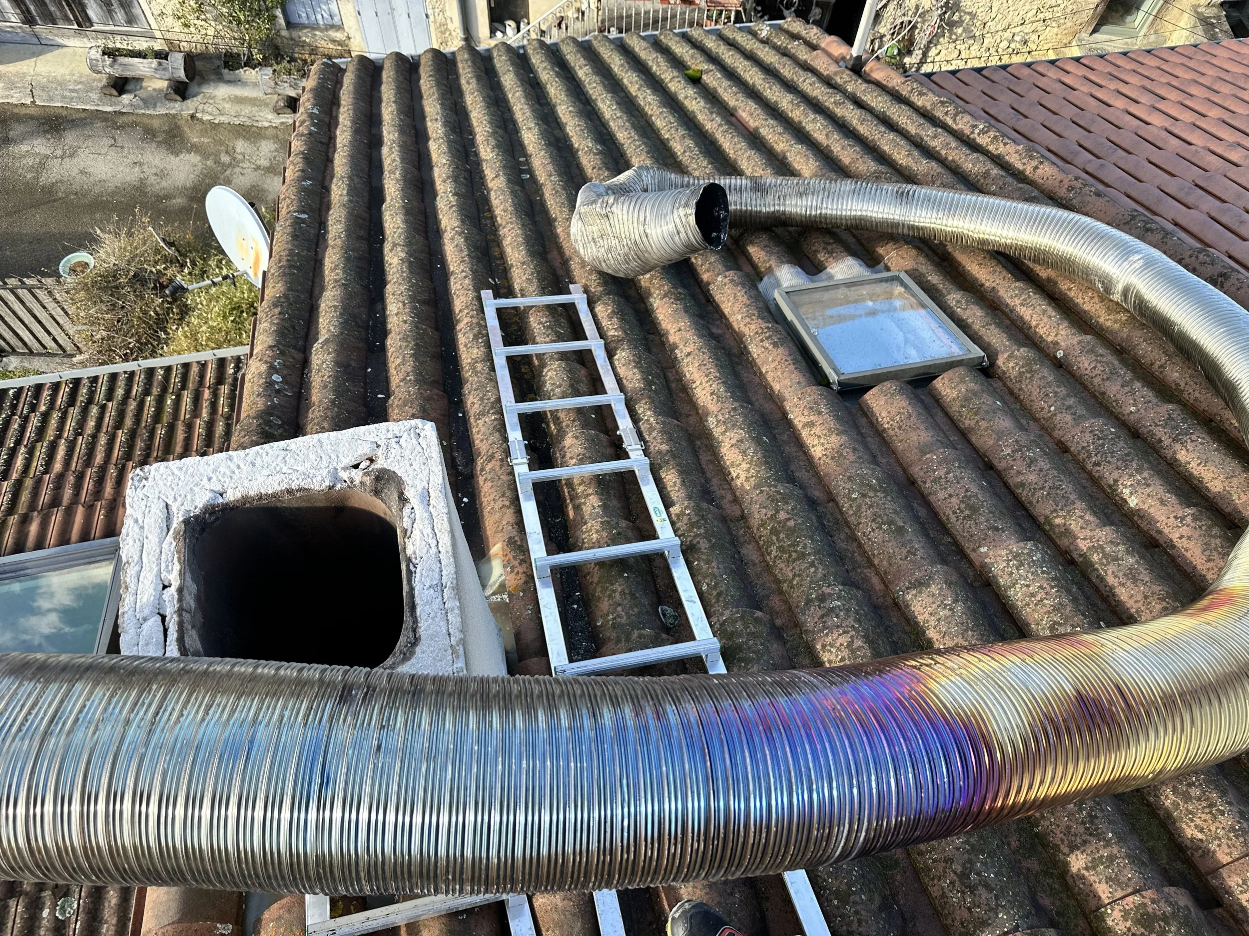 Vue d'un toit en tuiles avec un tube de ventilation en métal ondulé, une échelle pliante, et des éléments de ventilation en haut. À gauche, une fenêtre encadrée en pierre et une antenne satellite sont visibles.