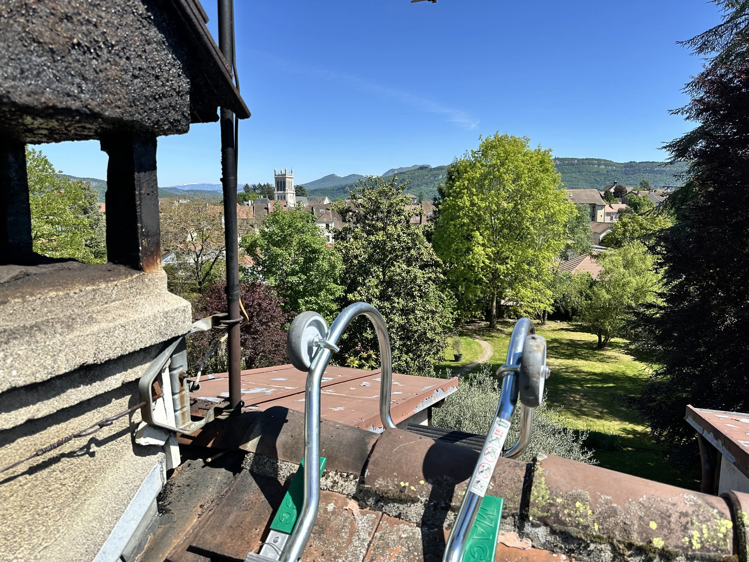 Vue d'un paysage urbain avec des bâtiments et une église, entouré de montagnes verdoyantes et un ciel clair. En premier plan, il y a une plateforme en bois avec des outils métalliques et un mur en pierre.