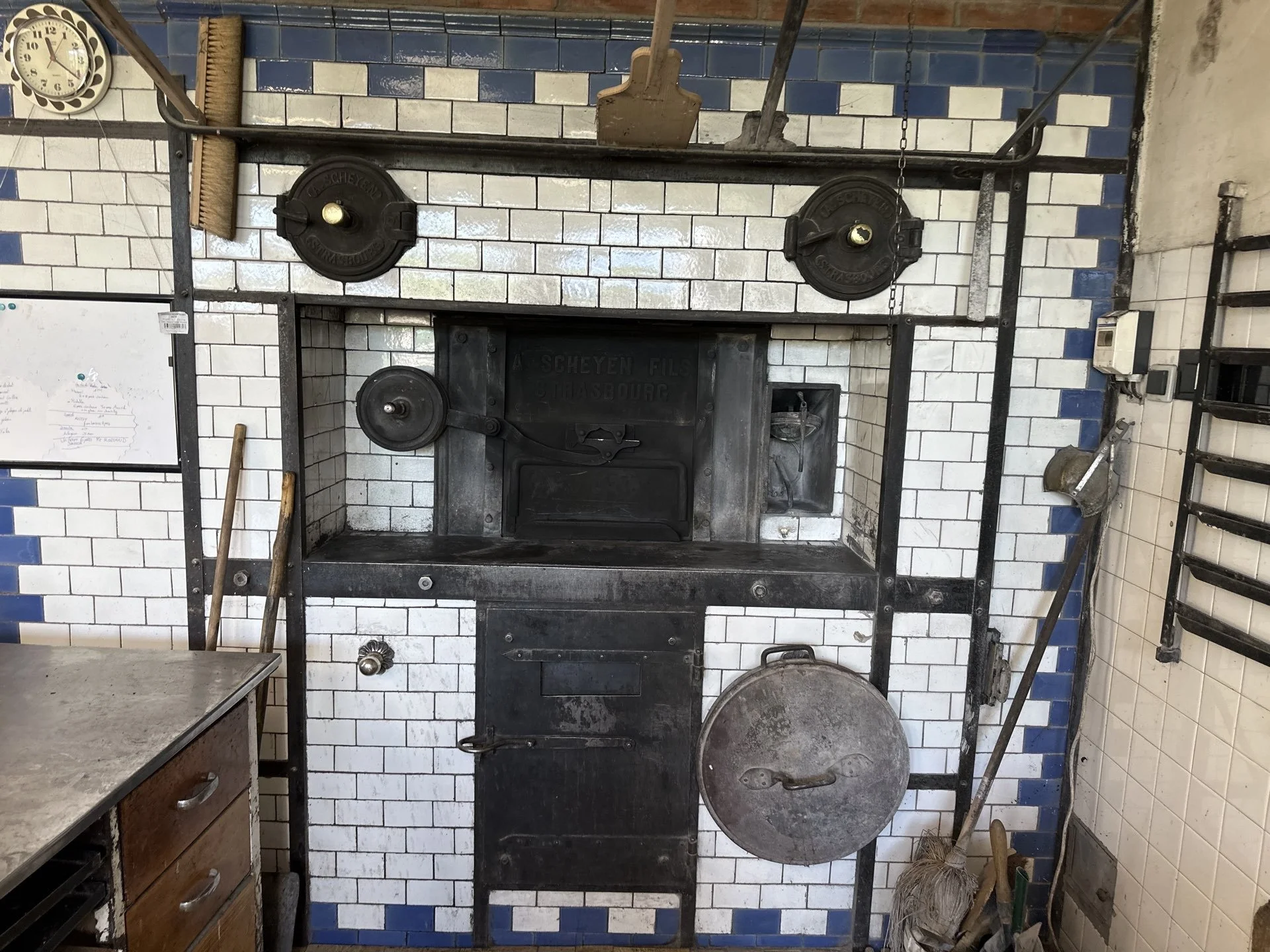 Mur en carreaux blancs et bleus avec une cheminée en fonte noire. Il y a des outils en métal autour, notamment une pelle à charbon, une brosse, et un seau. Un tableau blanc et un meuble en bois sont visibles à gauche. Le mur comporte également une horloge ronde, un thermostat, et un tapis de sol en bas à droite.