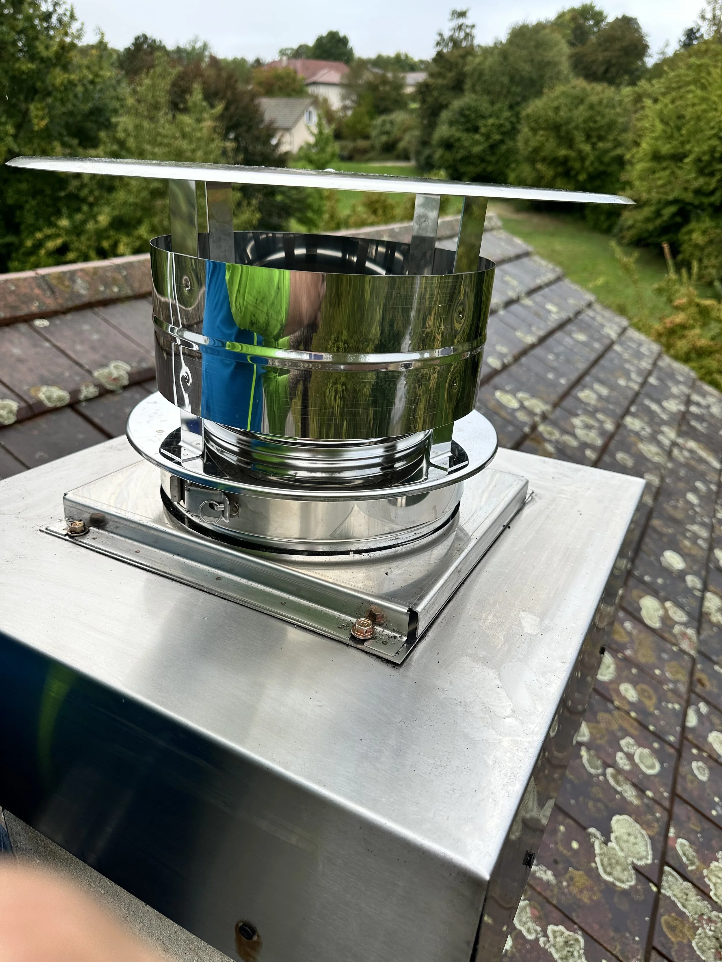 Une cheminée de toit en acier inoxydable installée sur un toit en tuiles, avec un environnement résidentiel et de nombreux arbres en arrière-plan.