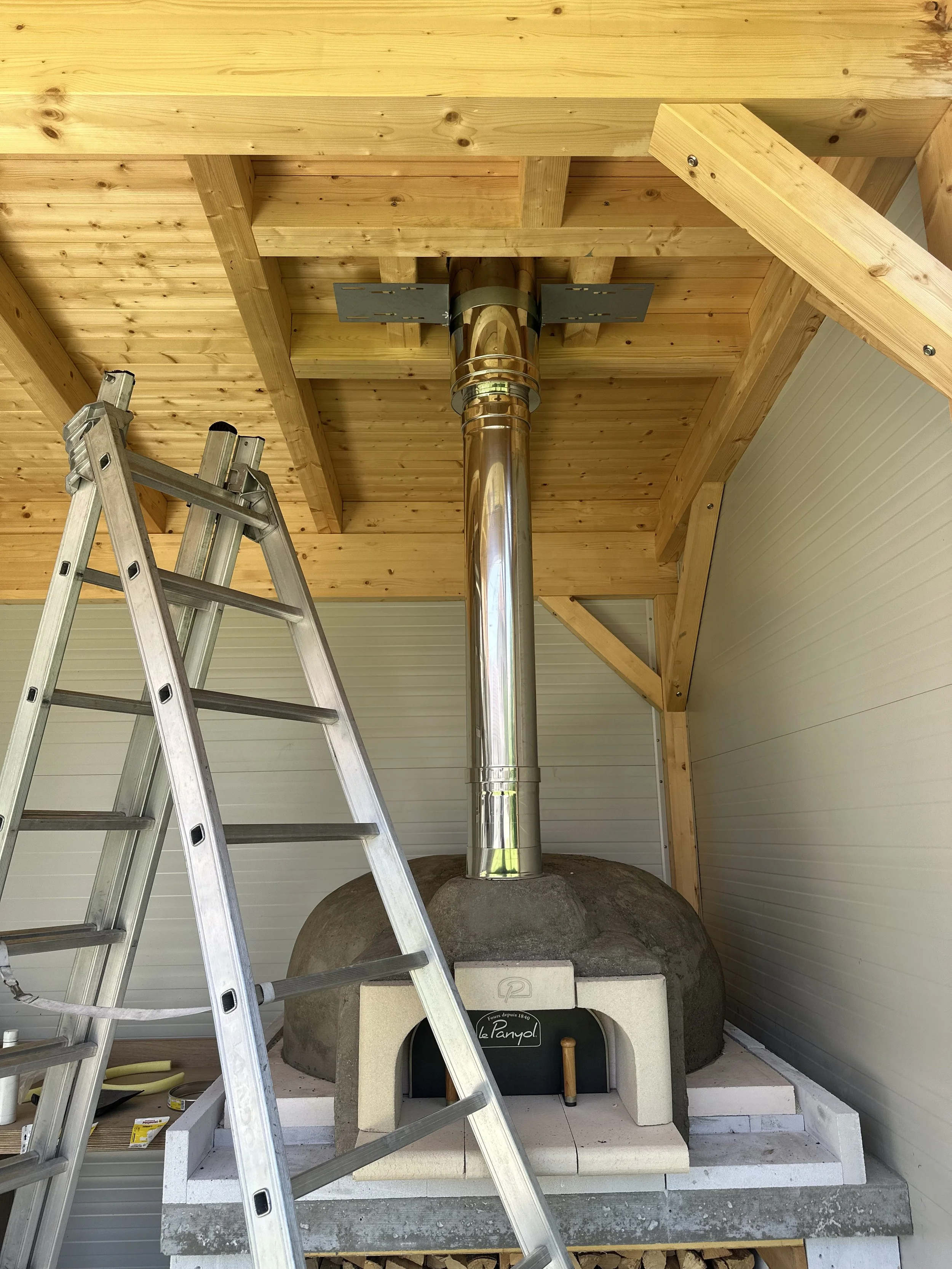 Cheminée en pierre avec conduit en métal, dans une structure en bois, avec un escabeau en aluminium à côté.