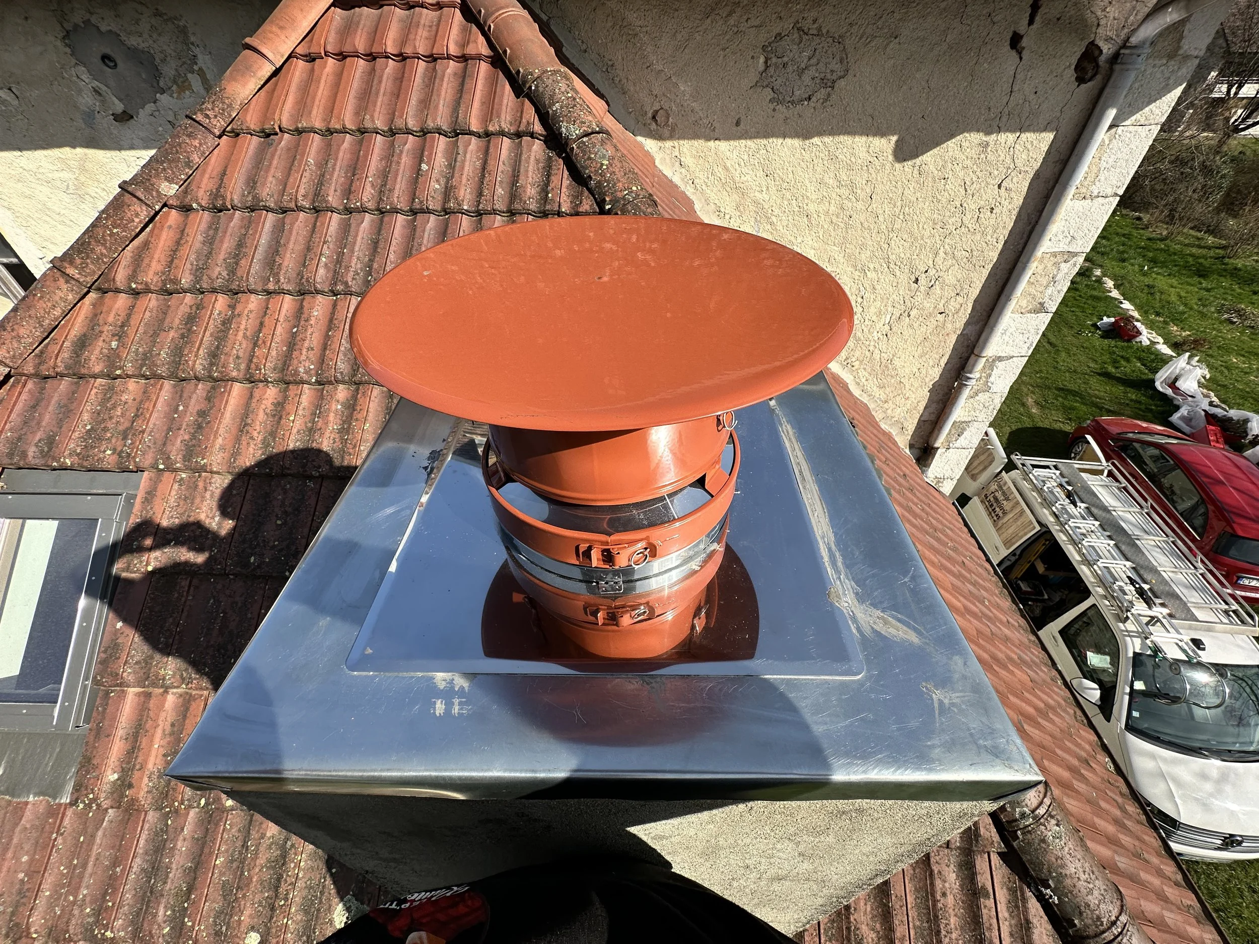 Une parabole orange installée sur un toit pluvial en tuiles rouges, avec un mur beige en pierre et plusieurs voitures garées en bas dans une rue.