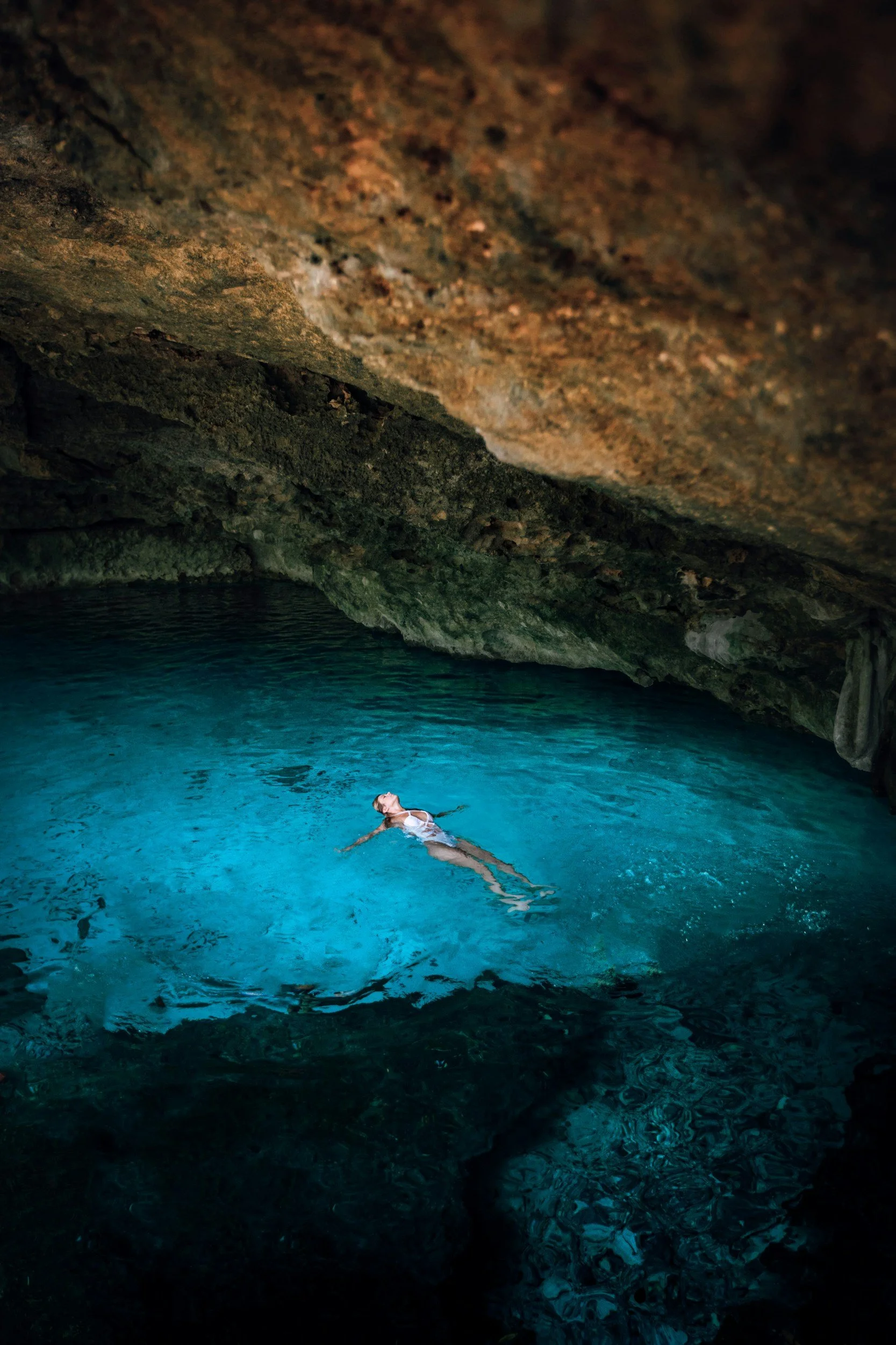 A woman in a swimsuit floating in clear blue water inside a dark cave with rocky walls.