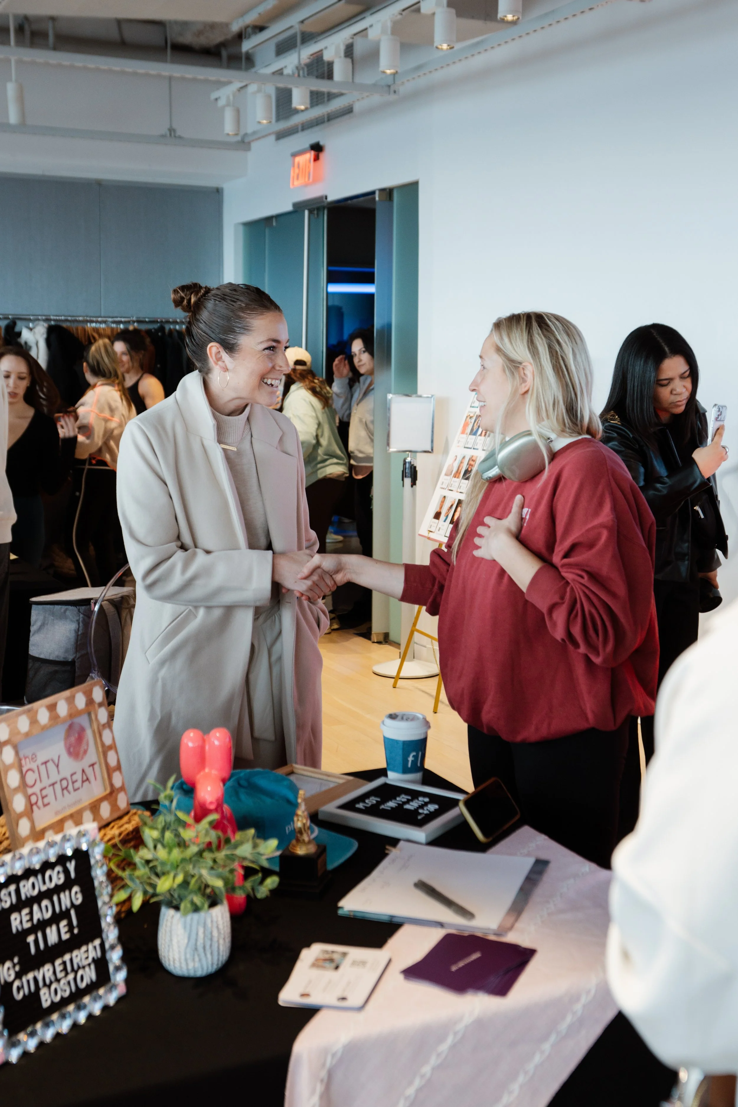 Two women shake hands at a booth during a networking event, with several people in the background and various display items on the table, including a sign with event details.
