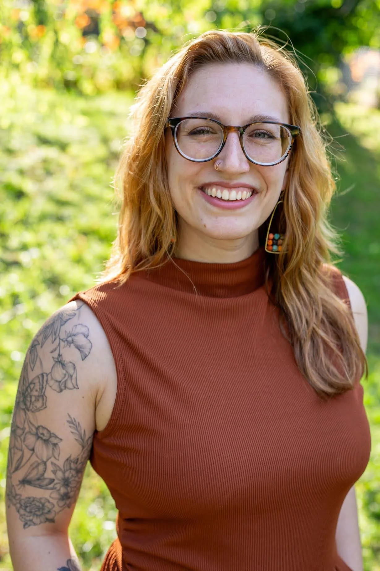 Close-up of a smiling woman with glasses, red hair, tattoos on her arm, wearing a sleeveless rust-colored top, outdoors with blurred green foliage background.