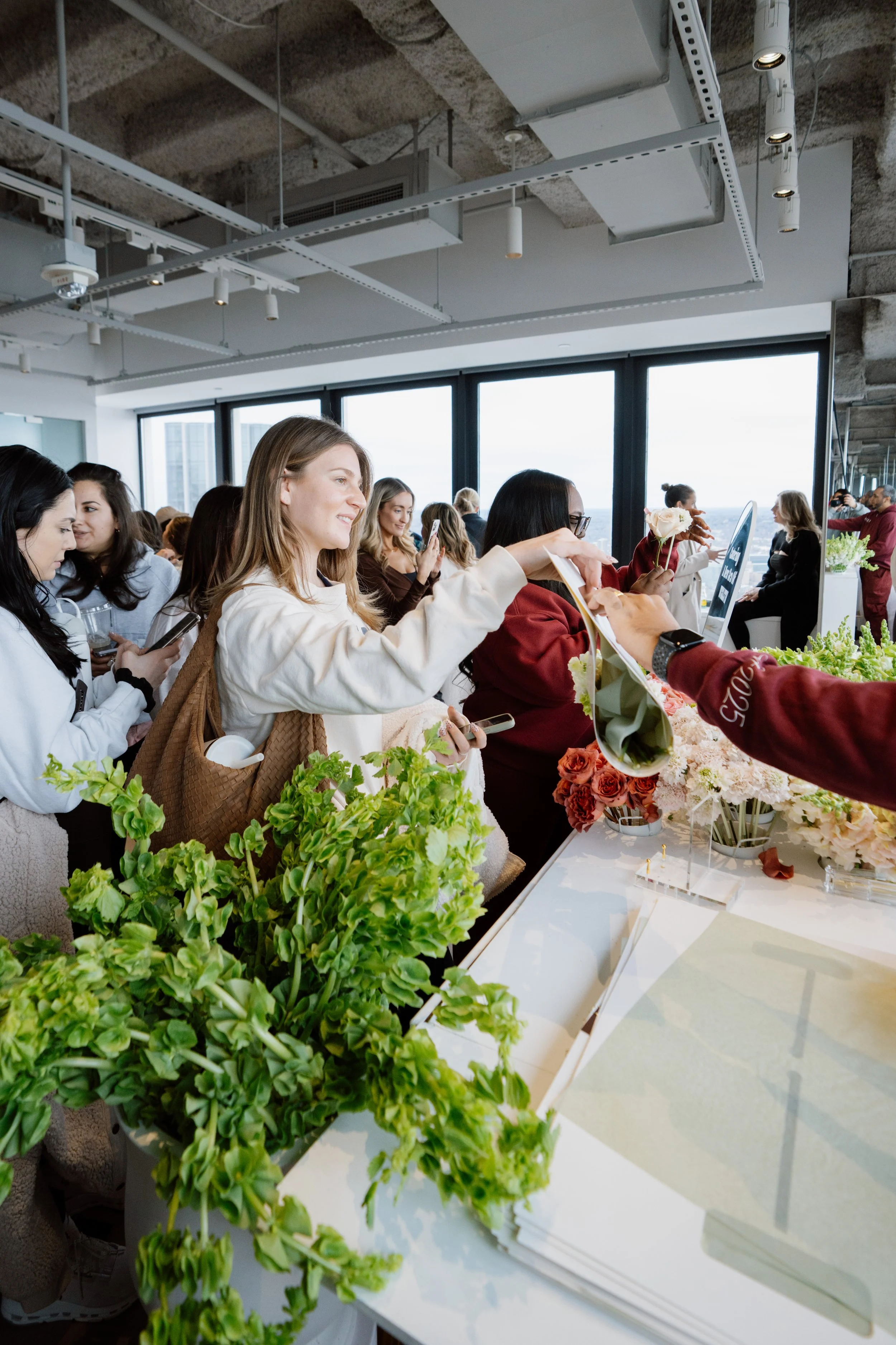 Many women gathered around a table near large windows, with some holding flowers and others looking at their phones or talking. The table is decorated with various flowers, including roses and green plants.