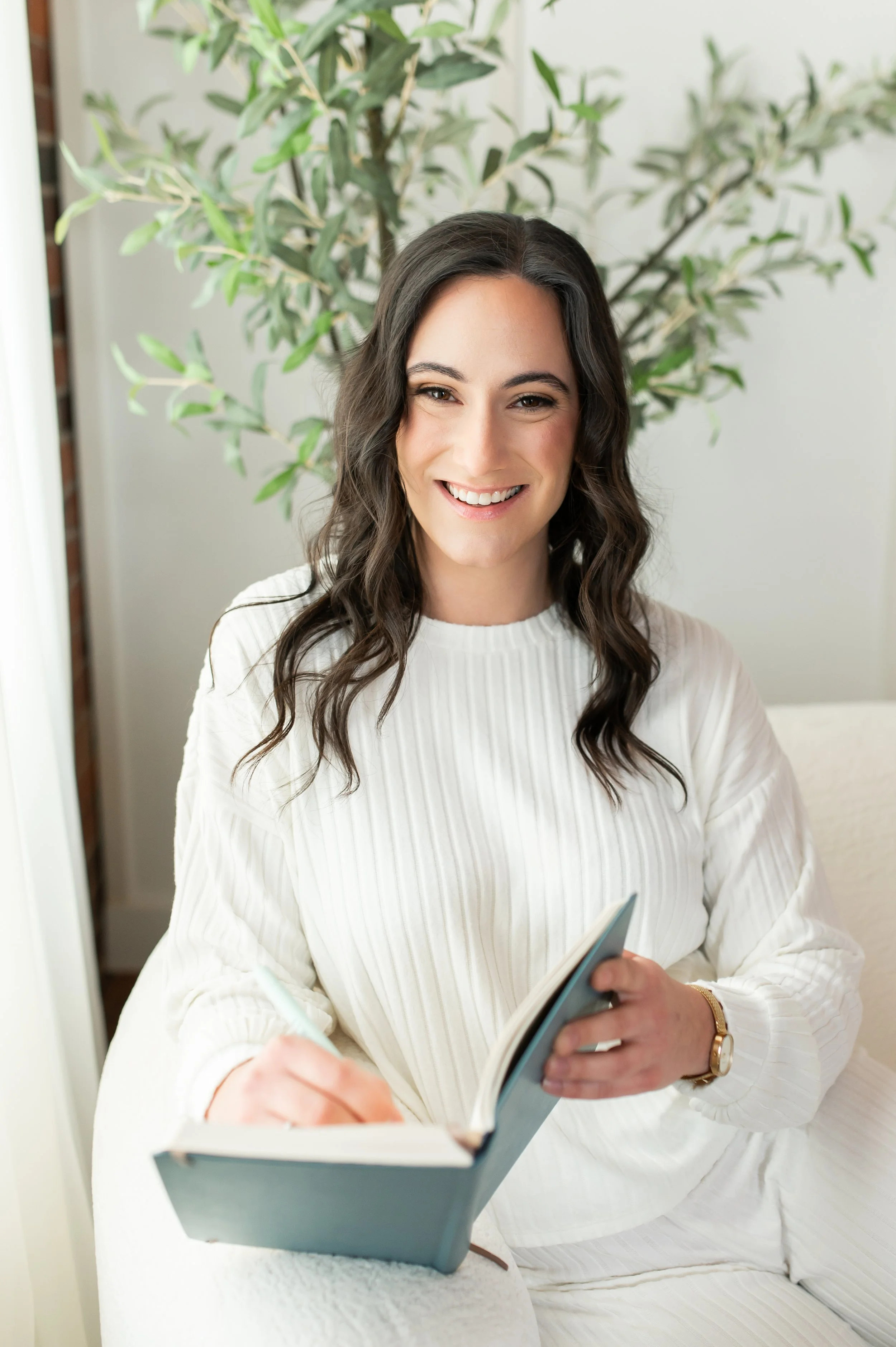 Smiling woman with wavy dark hair in a white top, sitting on a white couch with a green potted plant in the background, holding a blue book.