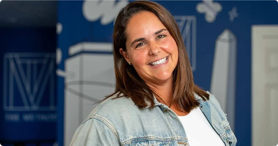 Smiling woman with shoulder-length brown hair wearing a denim jacket and white shirt, standing in front of a blue wall with white architectural graphics.