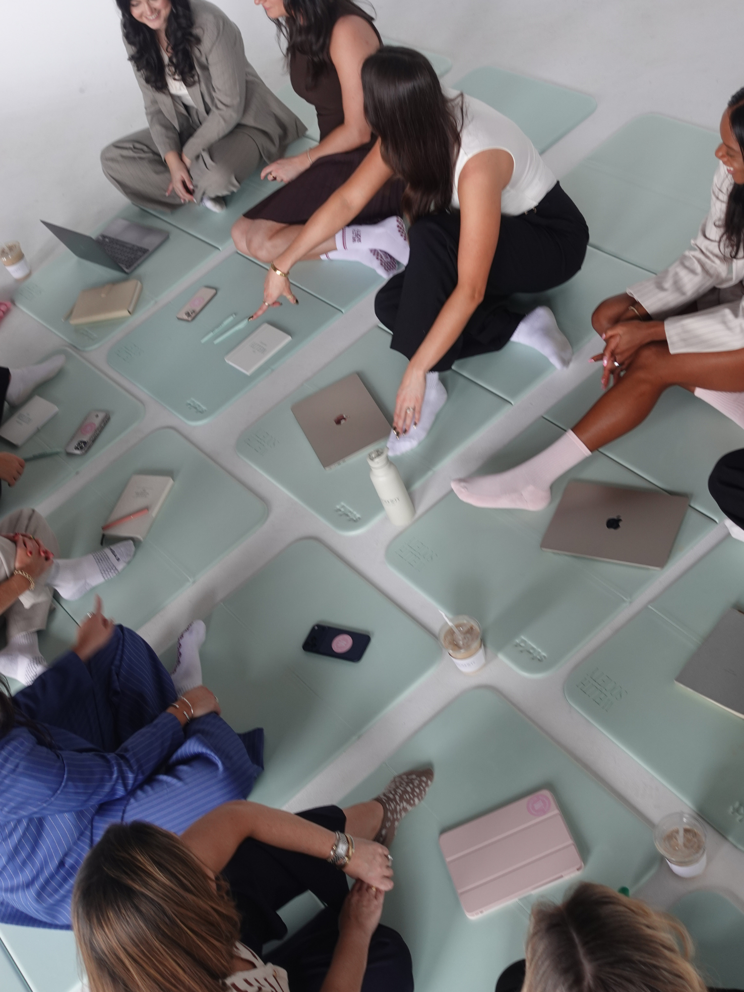 Group of women sitting on mint green benches in a circle, engaging in discussion, with notebooks, drinks, and phones on the benches, in a minimalistic room with white walls.