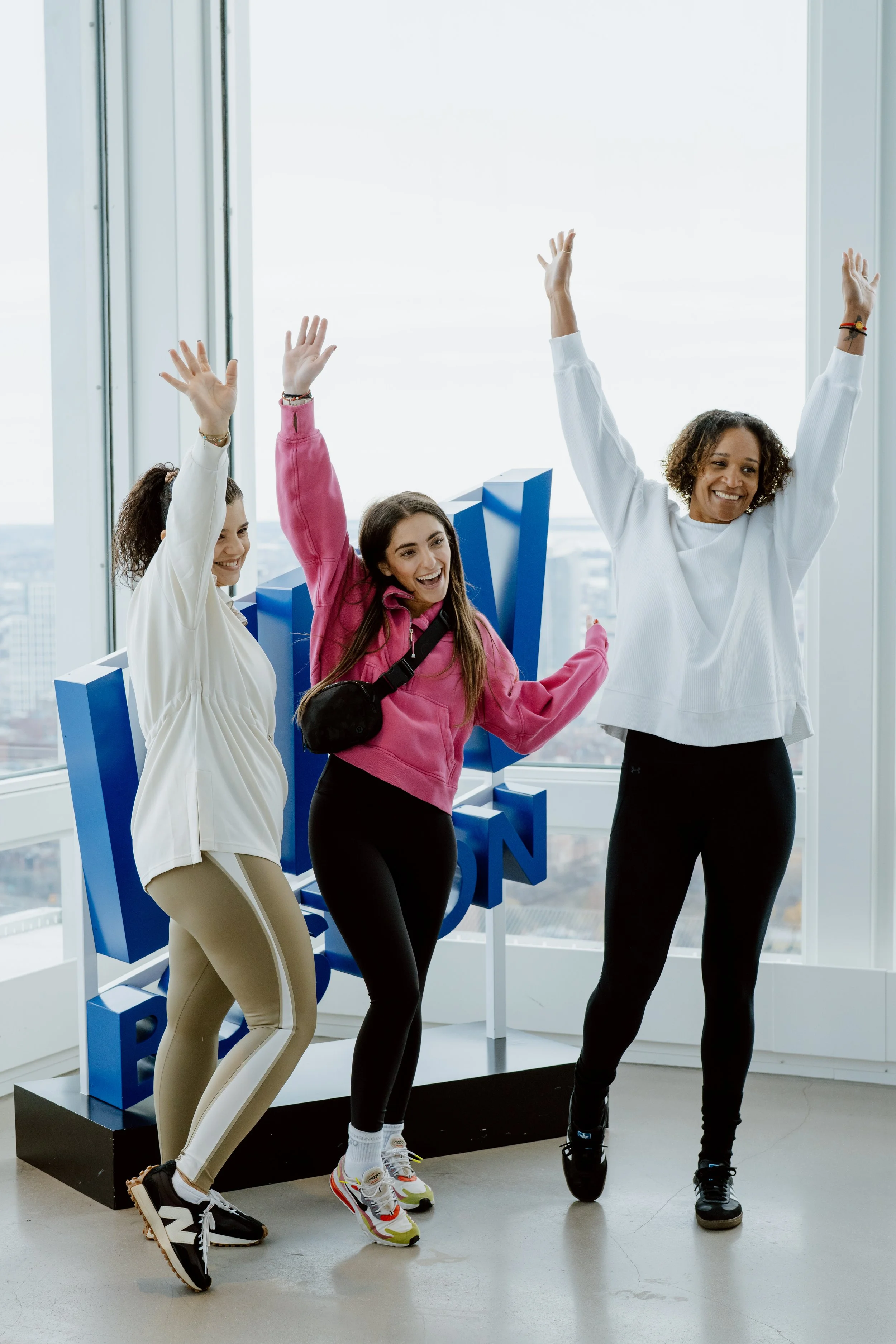 Four women are posing and raising their hands in front of a large window with a city view. One woman is leaning on a blue 3D sign that spells 'OPEN.' They are smiling and wearing casual, sporty clothing.