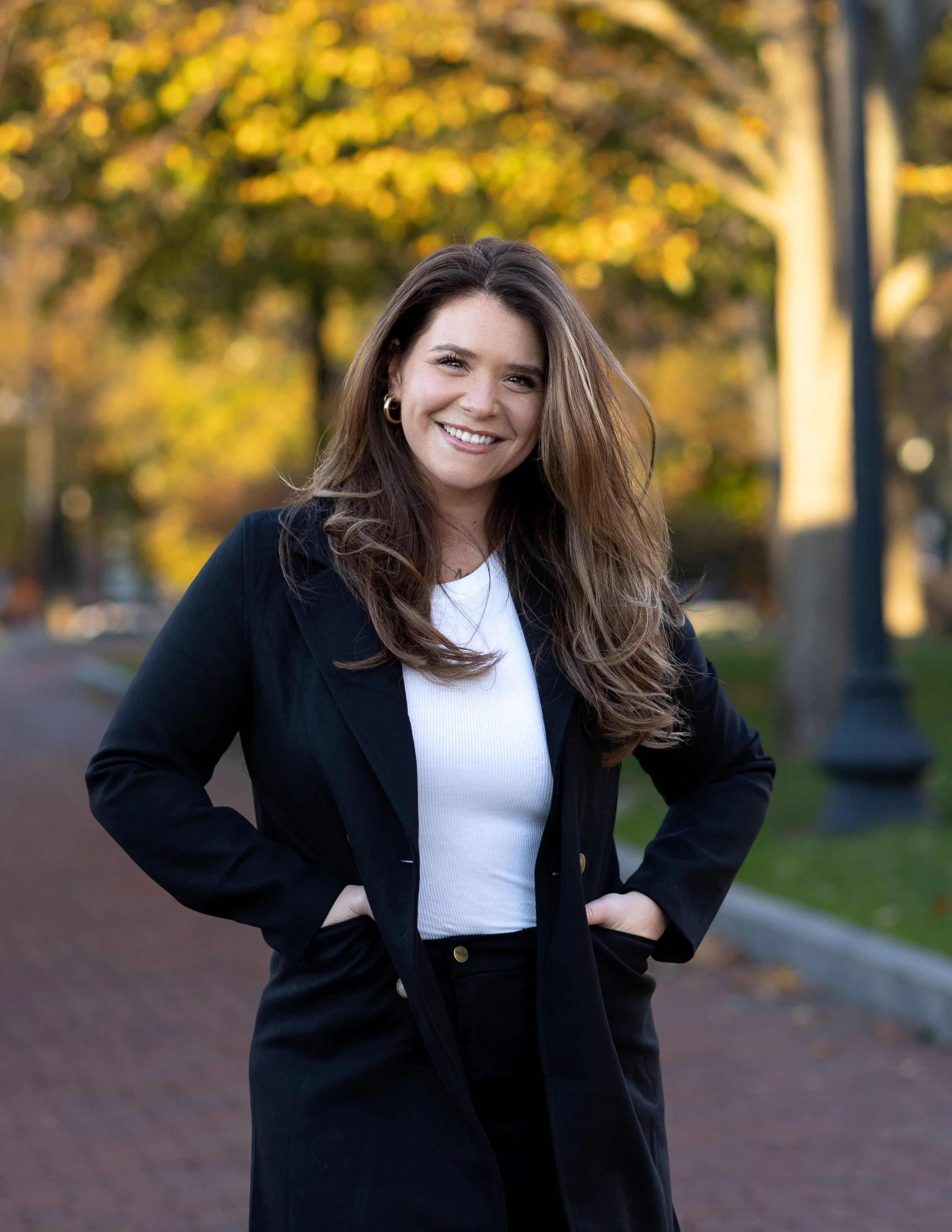 A woman with long brown hair smiling and standing outdoors in a park during fall, wearing a navy blazer over a white shirt with trees and golden leaves in the background.