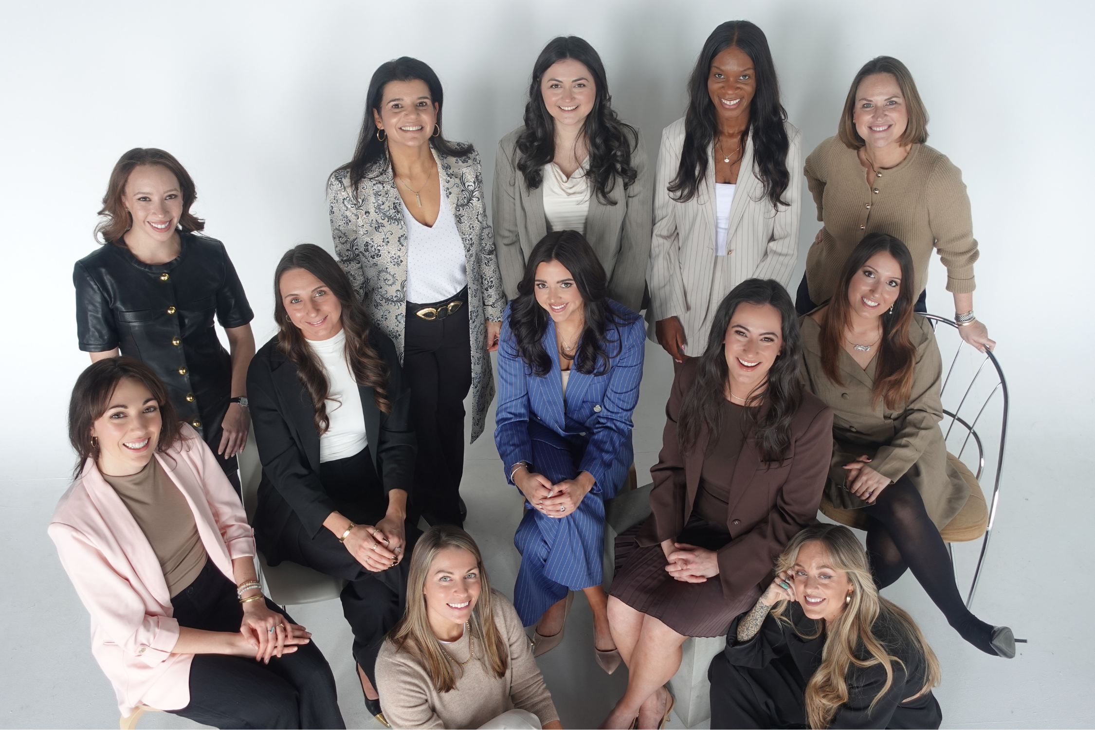 A diverse group of women gathered together for a photo in a studio setting, smiling and dressed in stylish business casual attire.