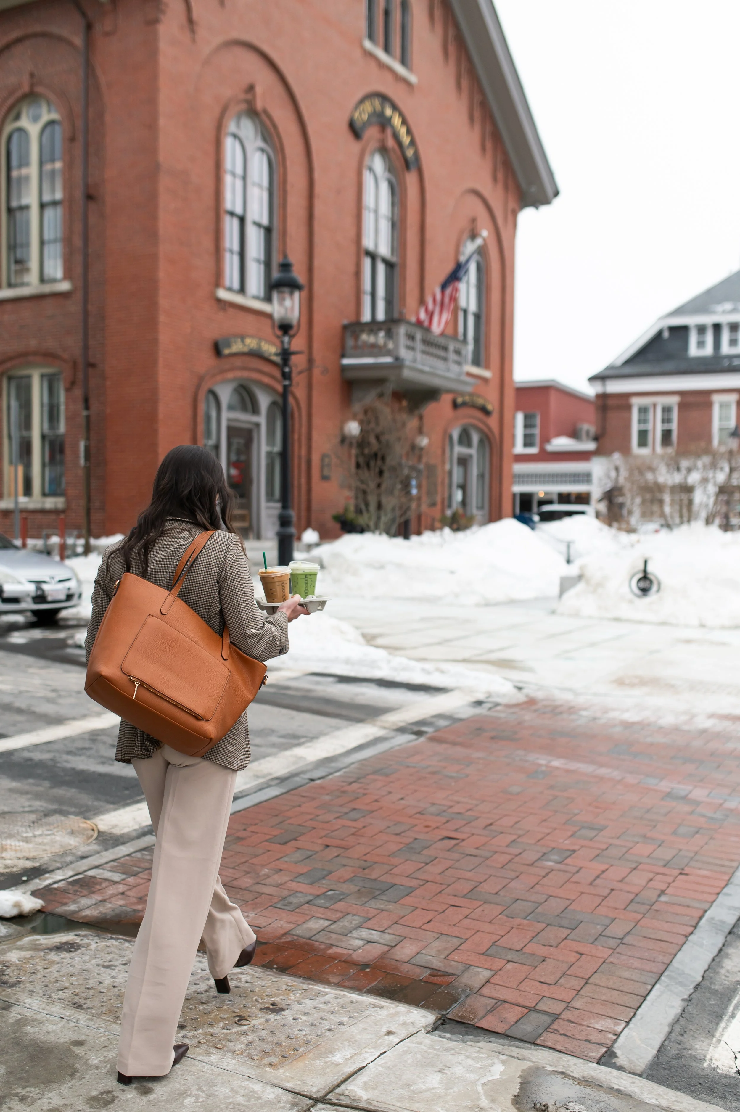 A woman with long dark hair, wearing a beige plaid blazer and cream-colored pants, carrying a large tan leather tote bag on her shoulder, walking on a brick sidewalk, holding two cups of coffee with green straws. There is snow on the ground and a red brick building with arched windows and an American flag on a small balcony behind her.