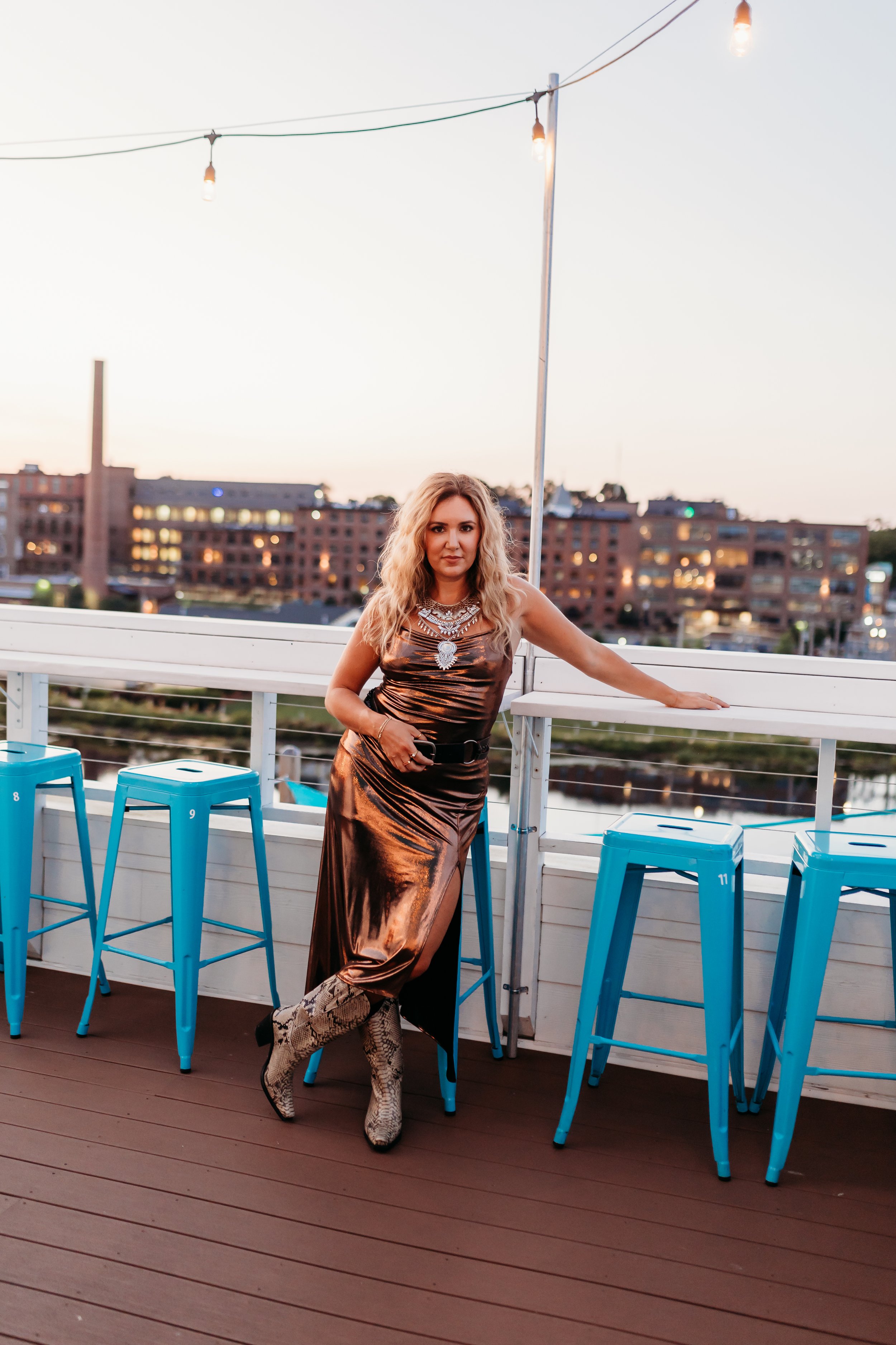 A woman with blonde curly hair stands on a rooftop patio at sunset, wearing a metallic brown dress, tall snakeskin-patterned boots, and large statement jewelry. She leans against a white railing with her right arm extended, with blue bar stools lined up in front of her and city buildings in the background.