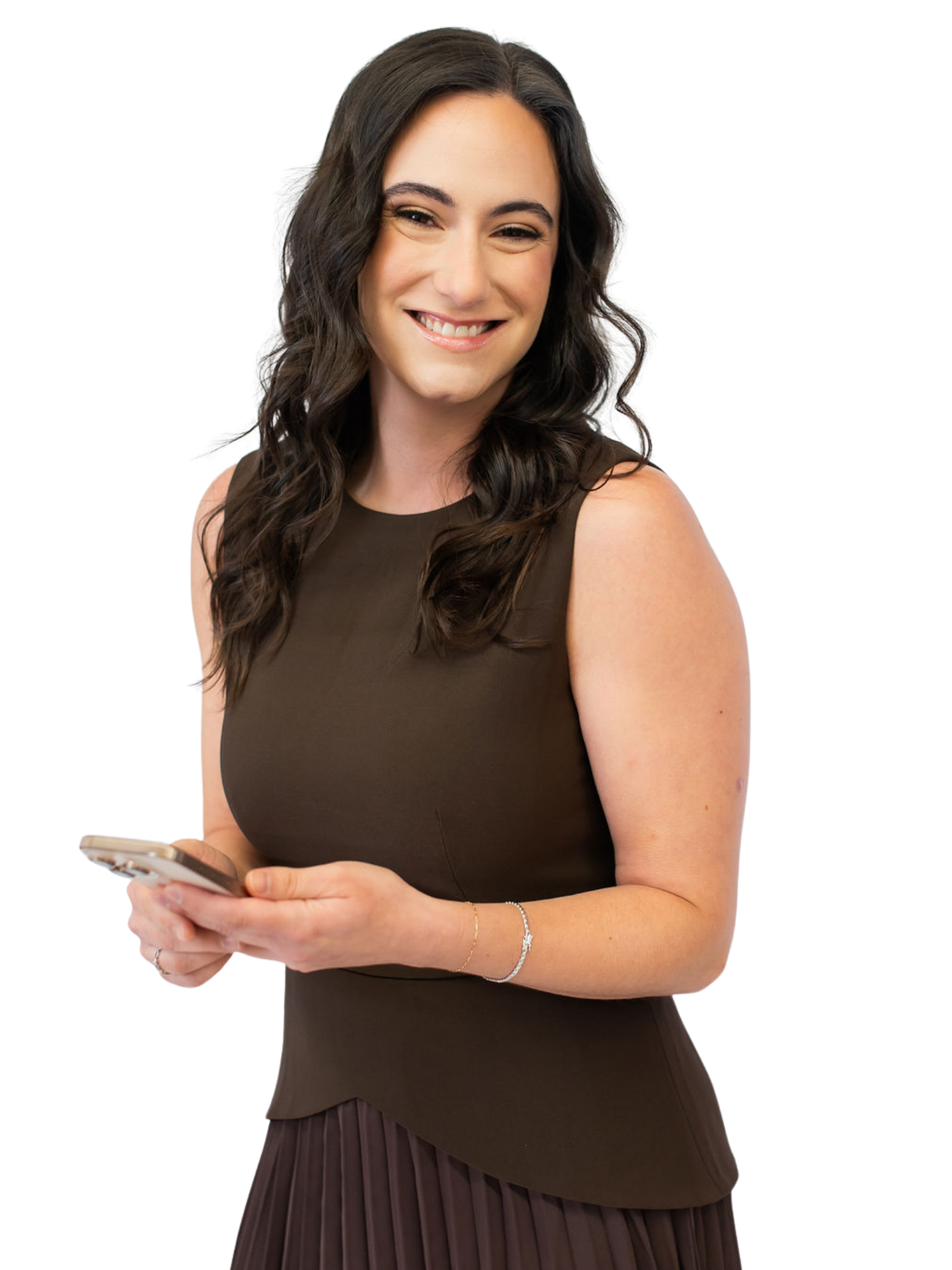 A woman with dark wavy hair wearing a dark sleeveless dress and a pleated skirt, holding a smartphone in her hands, smiling at the camera.