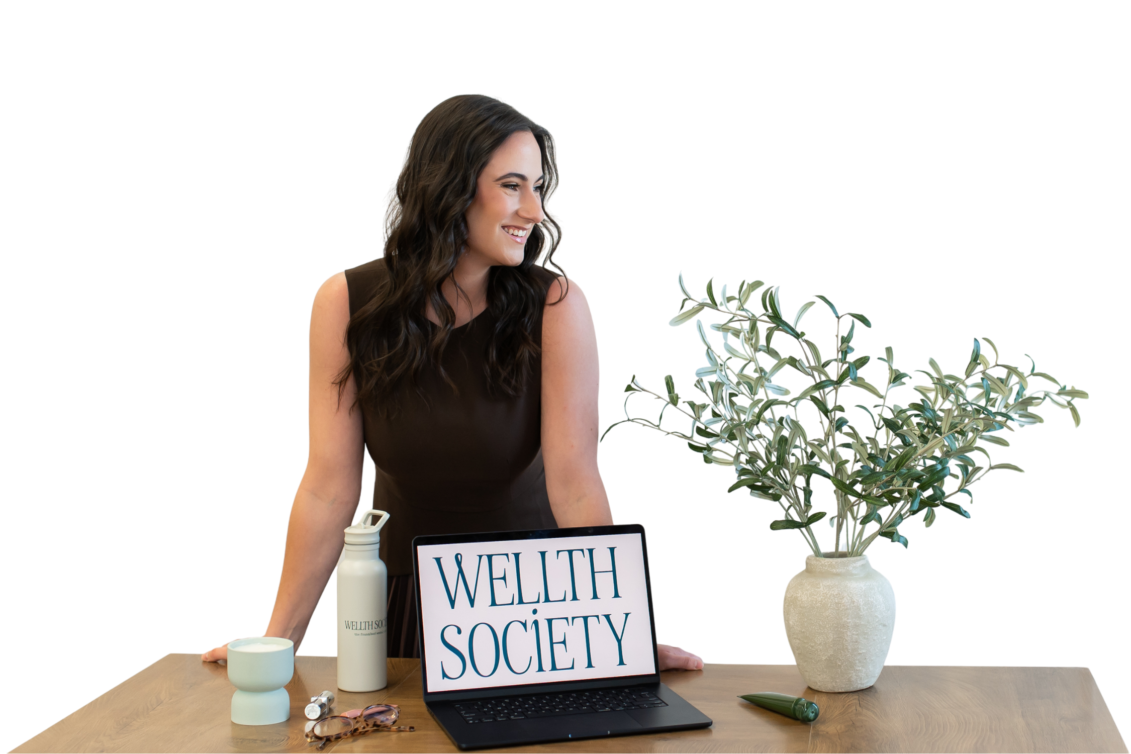 A woman with long wavy dark hair smiling, sitting at a desk with wellness products, a sign reading 'WELLTH SOCIETY,' a laptop, and green decorative items, including a large potted plant.